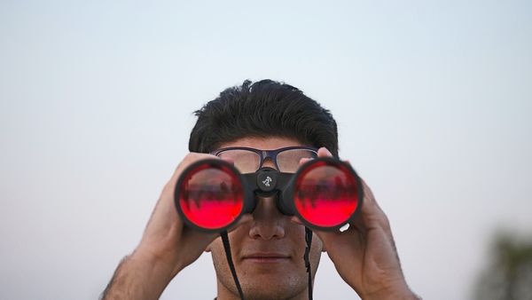 Ein Mann blickt durch ein großes Fernglas, auf den Gläsern spiegelt sich ein roter Himmel.