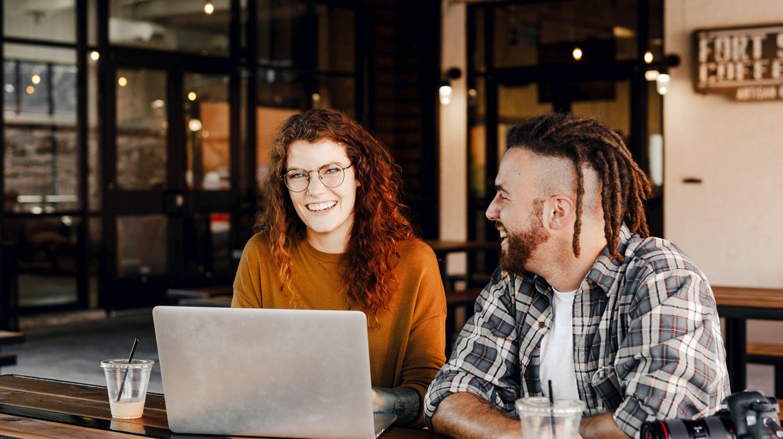 Eine junge Frau und ein junger Mann sitzen zusammen am Tisch und arbeiten an einem Laptop.