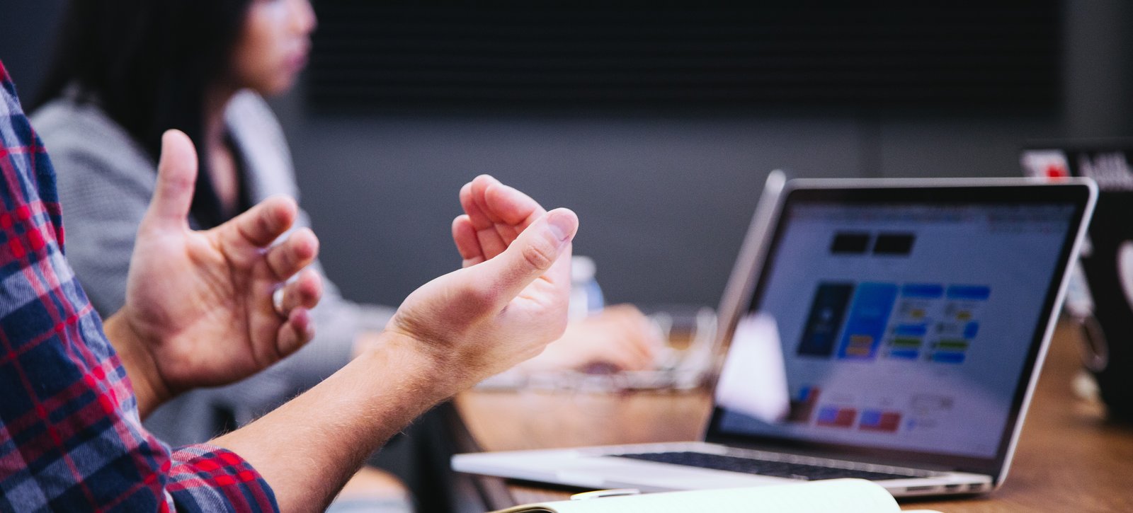 Forschung Ein Mann gestikuliert vor einem Notebook. Auf dem Tisch liegt ein Notizbuch und ein Smartphone.
