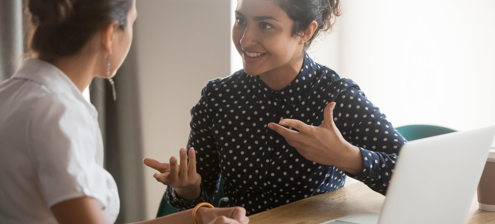 Weiterqualifizierung Zwei junge Frauen sitzen an einem Tisch mit einem Laptop und reden miteinander.