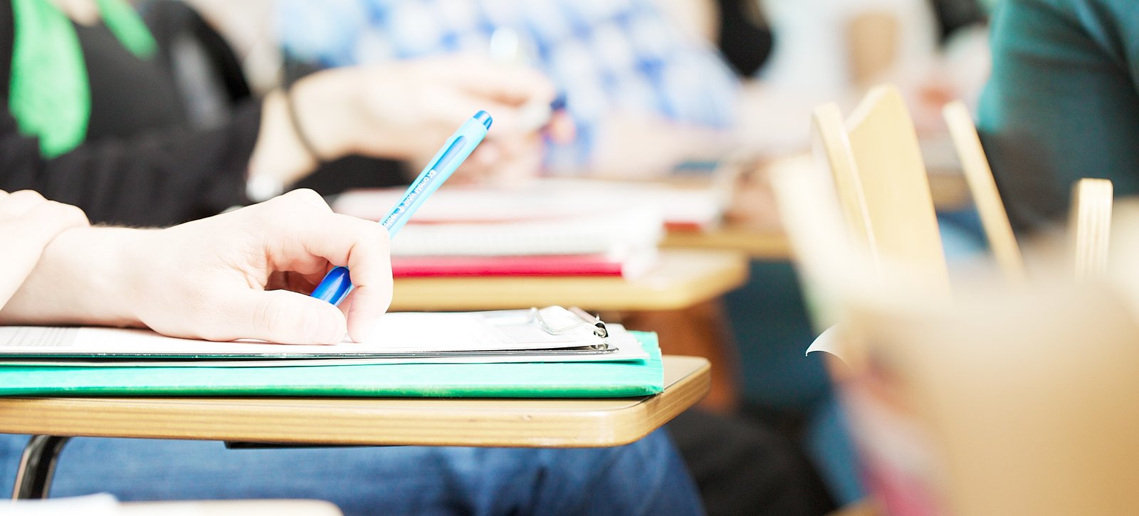 Rund ums Studium Blick auf einen Klapptisch mit Heften in einem Hörsaal. Studierende im Hintergrund.