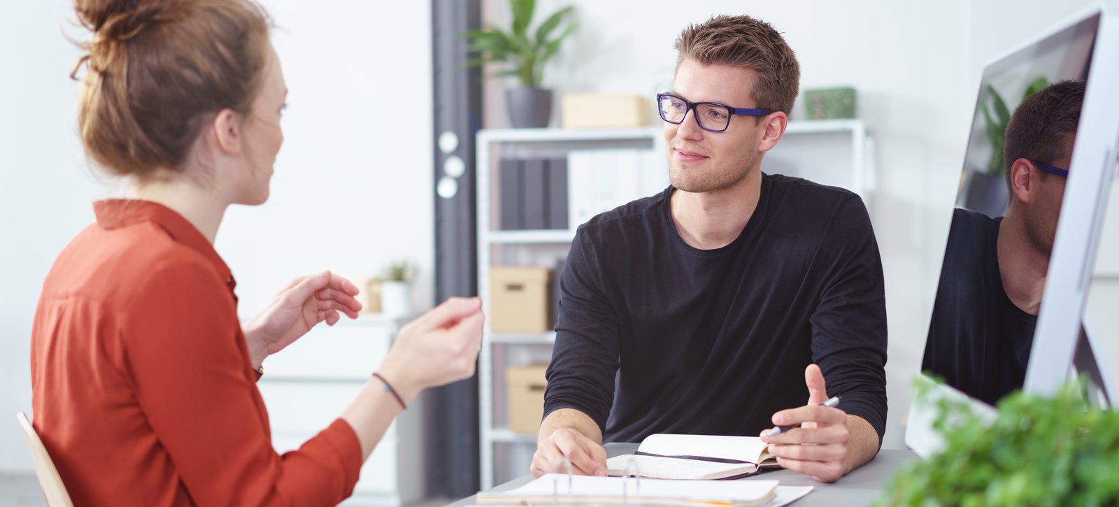 Studentische Beratung Eine junge Frau und ein junger Mann sitzen am Schreibtisch. Sie sprechen miteinander und beraten sich.